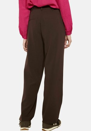 Person iført mørkebrune vidde bukser, lyserød ternet langærmet skjorte stukket ind og mørke sneakers, stående med ryggen til.