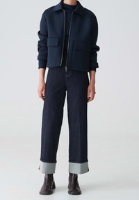 Giacca a cropped blu navy con chiusura a zip e due tasche frontali, abbinata a un paio di pantaloni in denim scuro a gamba larga con risvolti grigi. Stivali alla caviglia marroni.