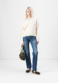 Beige gebreide trui, blauwe uitlopende jeans, zwarte sneakers met witte accenten en een olijfgroene tas. Achtergrond is effen wit.