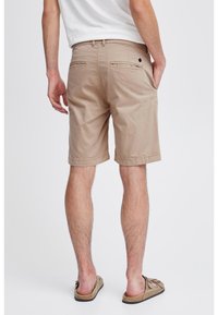Homme portant un short beige arrivant au genou, un t-shirt blanc et des sandales ouvertes couleur beige, se tenant dos à un fond blanc uni.