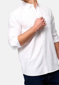 Chemise blanche à manches longues avec un poignet classique, présentant une poche poitrine. Le tissu semble lisse, avec un col classique et une coupe ajustée.