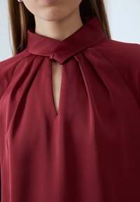 Blusa borgogna con design a pieghe, colletto alto e dettaglio a chiave sul collo, realizzata in tessuto morbido. La finitura testurizzata migliora l'aspetto.