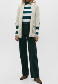 Femme portant un blazer beige, un col roulé rayé vert et blanc, un pantalon large vert foncé et des bottines marron à talons, posant devant un fond uni.