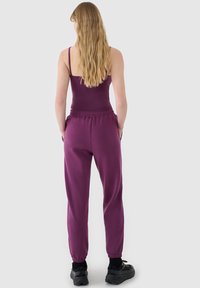 Femme aux longs cheveux blonds portant un débardeur violet et un pantalon de jogging assorti, debout les mains dans les poches, de dos.