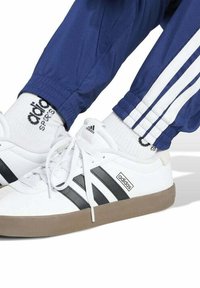 Zapatillas blancas con rayas negras, suelas beige y un acabado texturizado. Combinadas con pantalones de chándal azules con rayas laterales blancas.