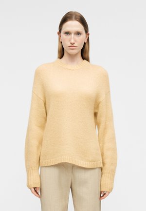 Kobieta z prostymi włosami, ubrana w luźny, jasnożółty sweter z dzianiny oraz beżowe spodnie, na tle jednolitego jasnego tła.