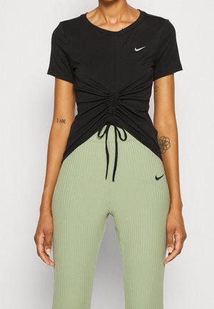 Kobieta w czarnym marszczonym topie Nike i jasnozielonych prążkowanych legginsach Nike z widocznymi tatuażami na ramionach.