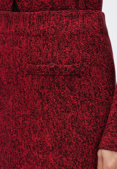 Textúrovaný pletený kabát v červenej a čiernej farbe, s bočným vreckom a dlhými rukávmi. Tkanina zobrazuje zmiešaný vzor farieb a stehov.
