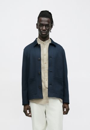 Mand med mørk hud og dreadlocks iført en marineblå jakke over en beige skjorte med knapper og hvide bukser, stående foran en hvid baggrund.