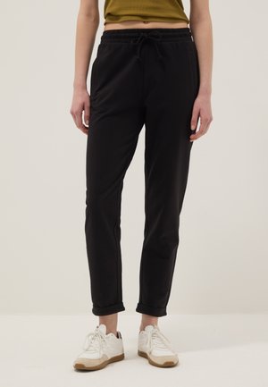 Mujer vestida con pantalones negros con cordón, combinados con zapatillas blancas y un top corto de color verde oliva, posando contra un fondo liso.