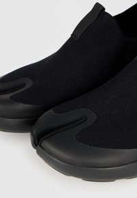 Μαύρα παπούτσια slip-on με σχεδιασμό διχαλωτού δακτύλου και πλεκτό επάνω μέρος, με ταινία στο μπροστινό μέρος για εύκολη εφαρμογή.