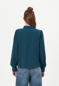 Teal blouse met lange mouwen en een kraag, gekenmerkt door geplooide schouderaccenten en een textuurstof. Gepaard met high-waisted vervaagde jeans.
