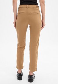 Person, der schmal geschnittene, beige Hosen mit Gesäßtaschen, schwarzen Gürtelschlaufen und schwarzen Plateauschuhen trägt, steht vor einem weißen Hintergrund.