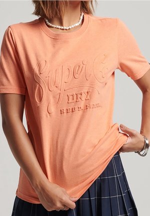 Kobieta ubrana w brzoskwiniowy t-shirt Superdry z wytłaczanym logo oraz granatową plisowaną spódnicę, ozdobiona perłowym naszyjnikiem i srebrnymi bransoletkami.