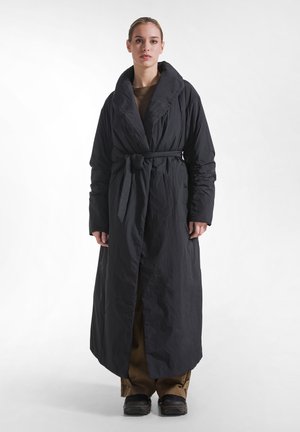 Donna con capelli biondi che indossa un lungo cappotto nero con cintura sopra pantaloni marroni e scarpe nere, in piedi su uno sfondo bianco.