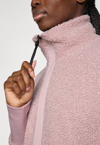 Nahaufnahme einer Person mit dunkler Haut, die den Reißverschluss einer pinken, strukturierten Fleecejacke mit hohem Kragen zieht.
