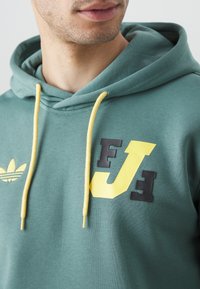 Grön hoodie med gula dragband, med en svart och gul grafisk design på bröstet och en broderad logotyp på vänster sida.