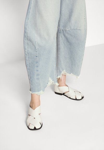 Sandali bianchi in pelle con design incrociato, suola piatta nera e punta aperta. Abbinati a jeans a gamba larga blu chiaro con orlo sfrangiato.