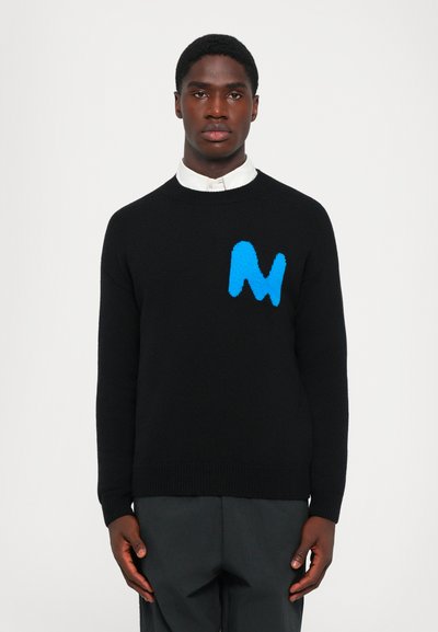 Joven vestido con un suéter negro con una "M" abstracta azul en el pecho y una camisa blanca con cuello debajo, de pie contra un fondo blanco.