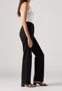 Jean noir taille haute avec une coupe droite et des poches arrière, associé à un débardeur blanc ajusté et des sandales noires à lanières.