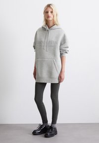 Hanorac oversized gri cu buzunar frontal și șnur, asortat cu leggings gri închis și pantofi negri cu șireturi pe un fundal simplu.