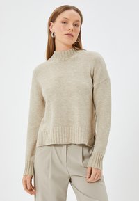 Beige neulepaita korkealla kauluksella, ribbaattu hihansuut ja helma. Kangas on pehmeä ja tekstuurinen, ja siinä on rento malli sekä alas pudotetut olkapäät.