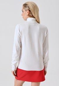 Frau mit nach hinten gegeltem blonden Haar, die einen weißen Rollkragenpullover und einen roten Rock mit goldenem Reißverschluss trägt, mit dem Rücken zu einer einfarbigen Hintergrund.