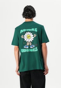 Zöld pamut póló, amelyen egy mosolygós arcú rajzolt százszorszép grafika látható, csillagokkal körülvéve, alatt pedig az "ADIDAS ORIGINALS" felirat található.