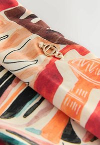 Farverigt stof med abstrakte mønstre i rød, orange, sort og beige, med en guldfarvet rund metal spænde med et geometrisk design.