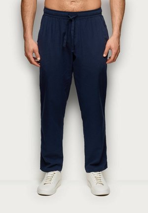 Mężczyzna w granatowych spodniach z wiązaniem i białych tenisówkach stoi na jednolitym jasnym tle.