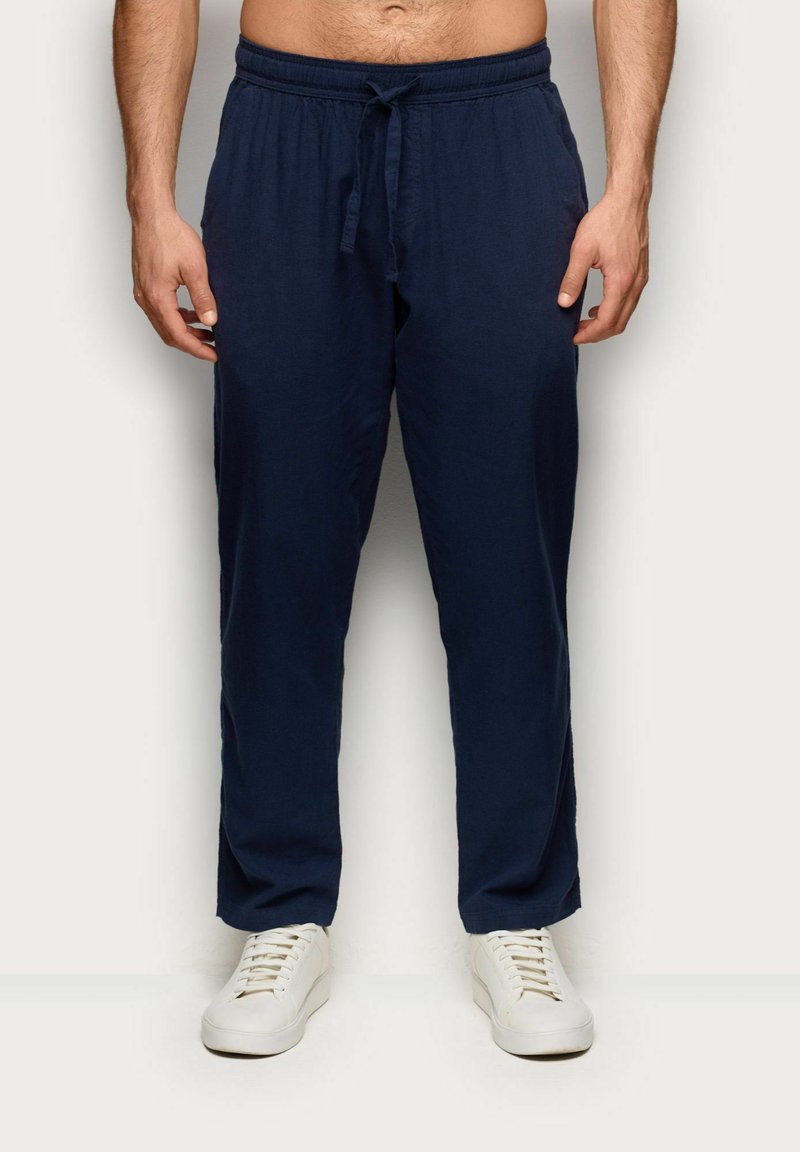 Hombre con pantalones de cordón azul marino y zapatillas blancas, de pie frente a un fondo liso y claro.