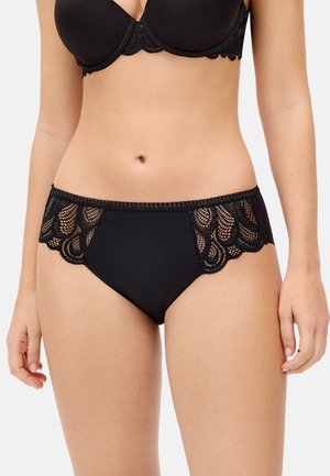 Mujer con braguitas negras con encaje y sujetador a juego, de pie contra un fondo claro y liso, con los brazos relajados a los lados.