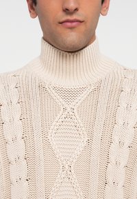 Hombre con un suéter de cuello alto de punto trenzado en color crema, recortado para mostrar la parte inferior de la cara y la parte superior del torso.