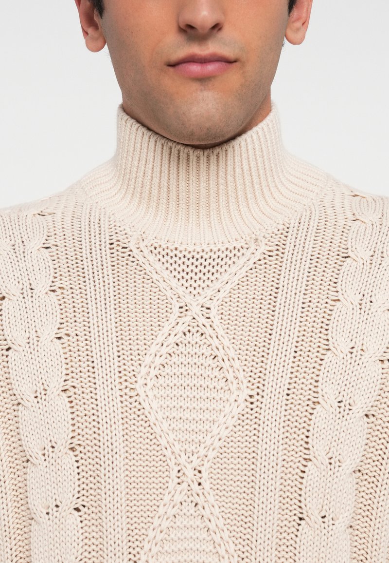 Hombre con un suéter de cuello alto de punto trenzado en color crema, recortado para mostrar la parte inferior de la cara y la parte superior del torso.