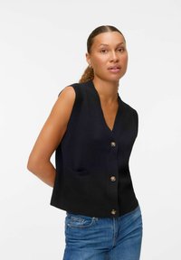 Črna brezrokavna cardigan jakna z V-izrezom, ki ima sprednji žep in zapenjanje na gumb z zaobljenimi robovi, v kombinaciji z modrimi kavbojkami.