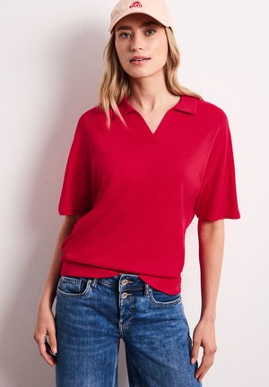 Kvinde iført en rød kortærmet poloshirt, blå jeans med dobbelt knaplukning og en lyserød cap med et rødt logo, stående mod en lys væg.