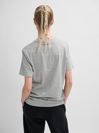 Persoon met drie gevlochten blonde paardenstaarten, draagt gouden oorringen, een grijs t-shirt en zwarte broek, staat met handen in de zakken.