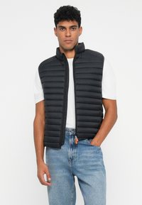 Sort, puffy, ærmeløs vest med hævede syninger og en høj krave. Båret over en hvid t-shirt, paret med blå jeans. Orange lynlås træksnor detalje.
