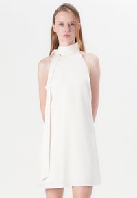 Baltas bezpiedurkņu kleita ar augstu apkakli un pie kakla sasietu akcentu. Gluda auduma un relaksēta piegriezuma, līdz ceļgaliem.