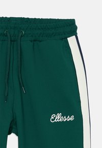 Zelené športové tepláky s elastickým pásom, šnúrkou na stiahnutie, bočnými pruhmi v bielej a námorníckej farbe a vyšitým logom "Ellesse" v bielej farbe.