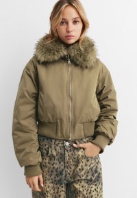 Kratka olivno-zelena bomberska jakna z umetnim krznom na ovratniku, rebrastimi manšetami in zapiranjem na zadrgo, v kombinaciji s jeans hlačami s pikami.