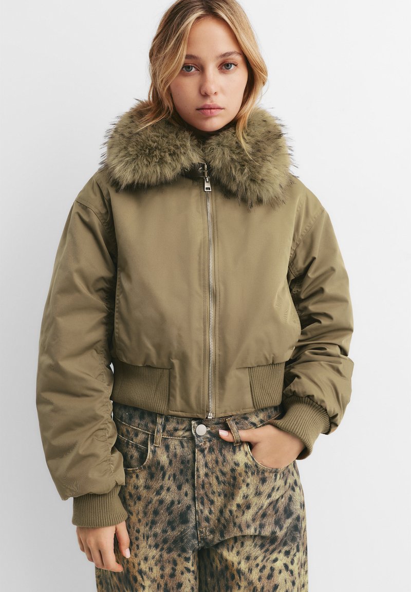 Kratka olivno-zelena bomberska jakna z umetnim krznom na ovratniku, rebrastimi manšetami in zapiranjem na zadrgo, v kombinaciji s jeans hlačami s pikami.