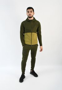 Chándal con capucha verde de diseño bicolor; cuenta con un cierre frontal, bolsillos laterales y pantalones ajustados. Zapatillas negras completan el look.