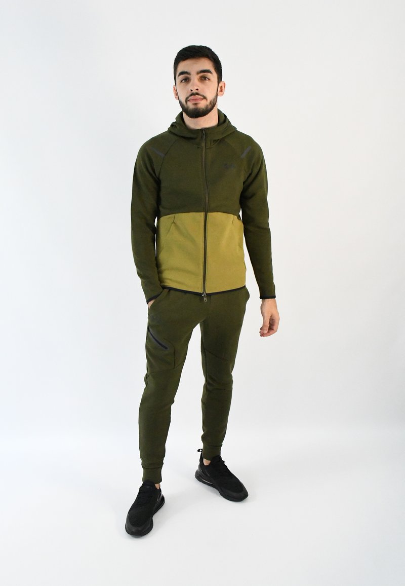 Chándal con capucha verde de diseño bicolor; cuenta con un cierre frontal, bolsillos laterales y pantalones ajustados. Zapatillas negras completan el look.
