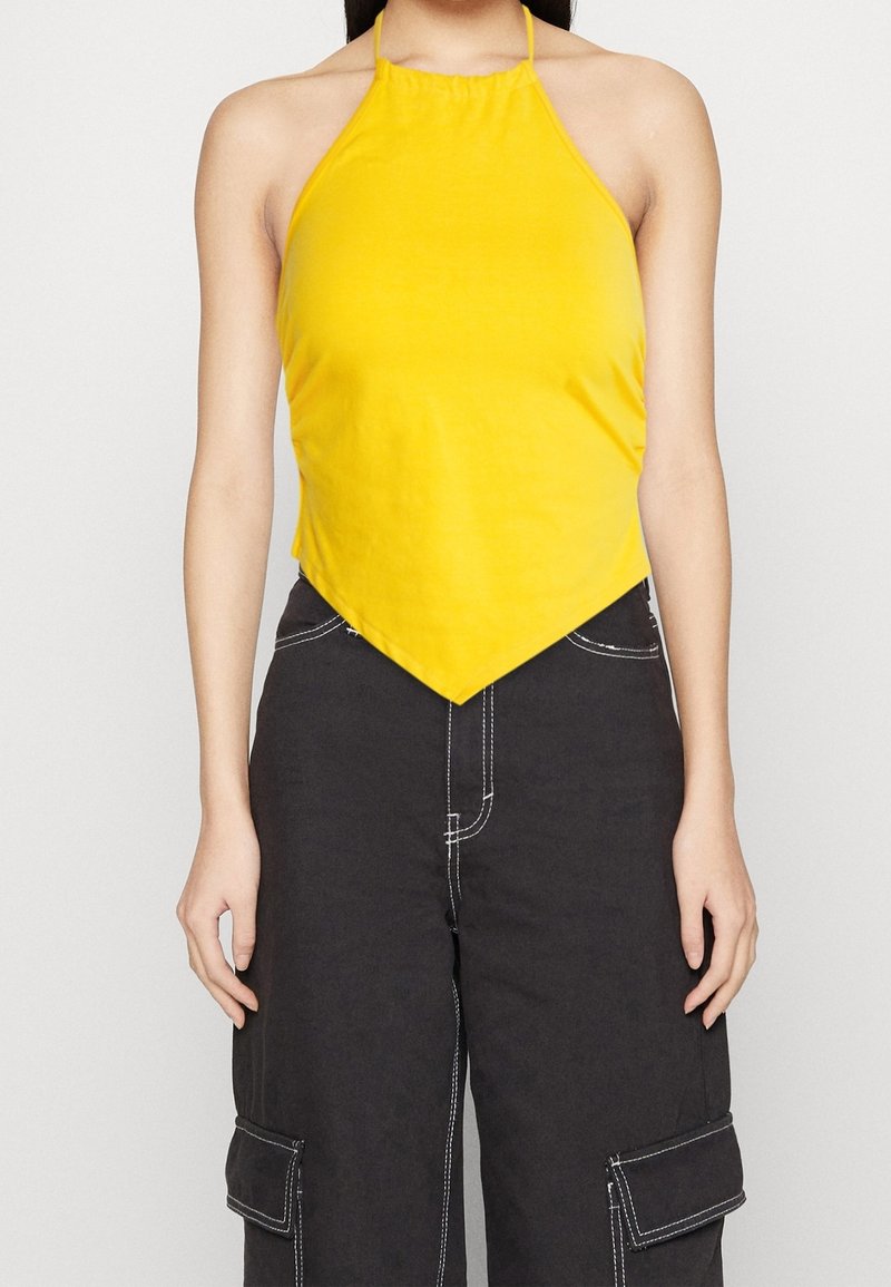 Woman wearing a sleeveless yellow halter neck top with a pointed hem paired with black cargo pants featuring white stitching.