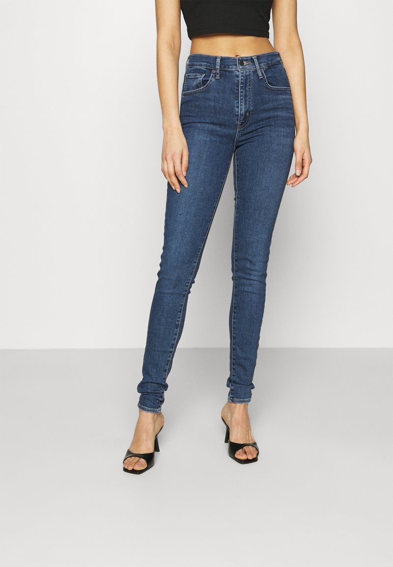 Femme portant un jean skinny bleu taille haute, un crop top noir et des sandales noires à talons ouverts, debout devant un fond uni.