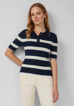 Frau trägt ein dunkelblau-weiß horizontal gestreiftes Polo mit kurzen Ärmeln und hellbeige Hose, steht mit einer Hand in der Hüfte.