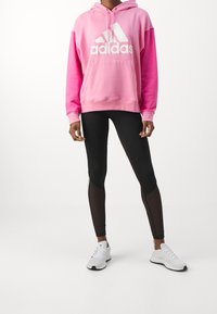 Rosa huvtröja med en vit Adidas-logotyp, i kombination med svarta leggings med genomskinliga paneler. Modellen bär vita sportskor.