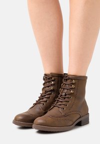 Botas de tobillo de cuero marrón con acabado texturizado, cierre de cordones en la parte delantera, ojales de metal y detalles de brogue en la puntera. Tacón bajo y cuadrado.