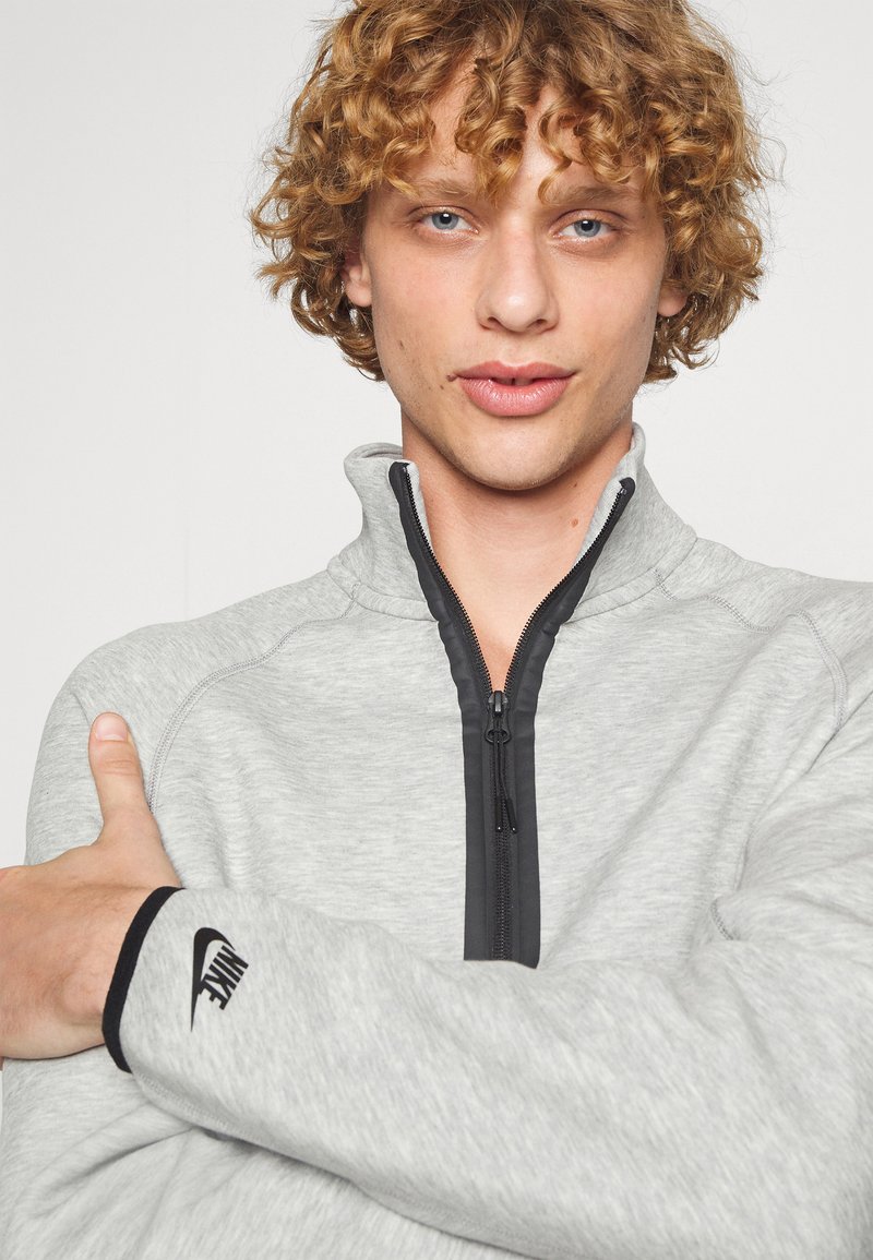 Sweat-shirt zippé gris avec des accents noirs et le logo Nike sur la manche. Doté d'un col montant et d'un tissu texturé.